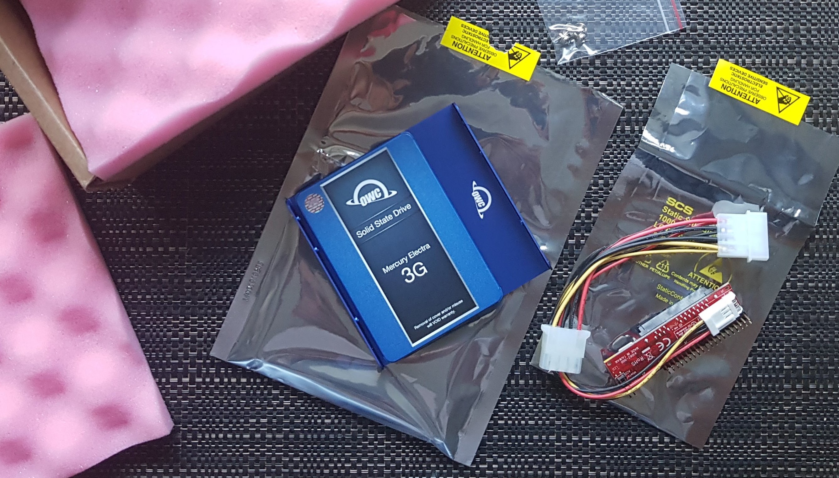 A solid-state hard drive, surrounded by a box, some static-shielding packaging, and a EIDE to SATA adapter